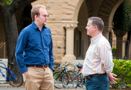 Two male Stanford affiliates talking in the quad, arches in background.