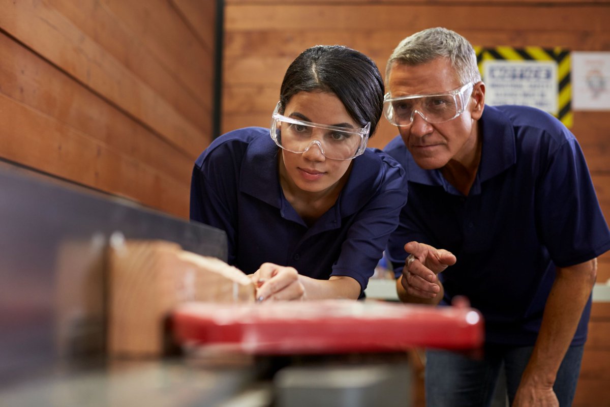 Older gentleman showing young lady how to use a bladed tool, both wearing goggles