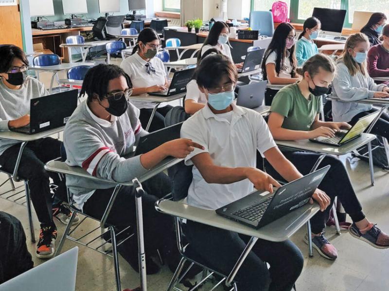 Masked students work on laptops in a classroom. 