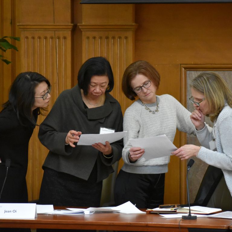 Four women standing and examining printed papers next to a conference room table with scattered paper on it