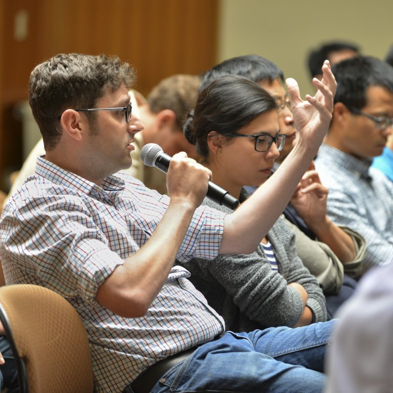 Event attendee seated in a lecture hall among other audience members, speaking to microphone. 