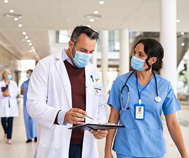 Two masked medical staff confer in a hospital corridor.